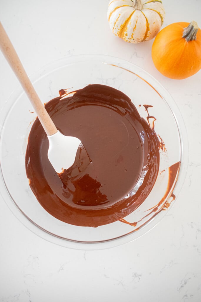 glass mixing bowl with melted chocolate and a spatula on a marble counter with small pumpkins.