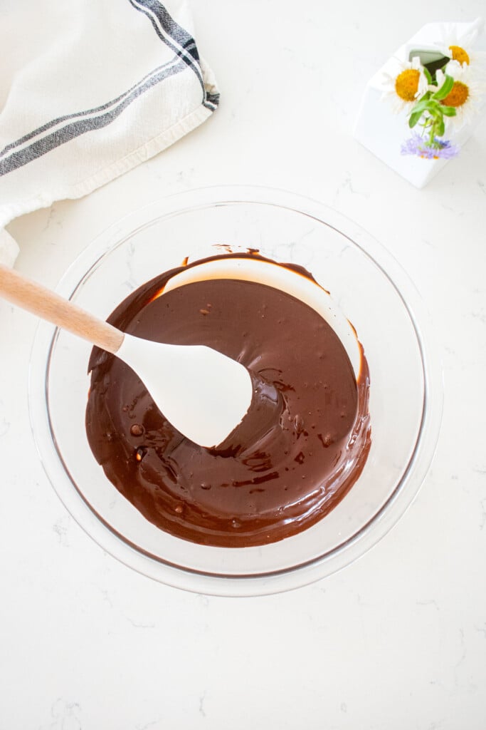 glass bowl with melted chocolate and a spatula on a marble counter with a small vase of flowers.