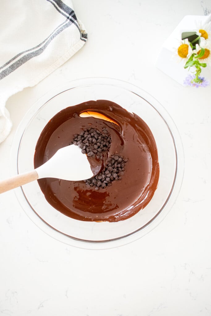 glass bowl with melted chocolate, chocolate chips, and a spatula on a marble counter with a small vase of flowers.