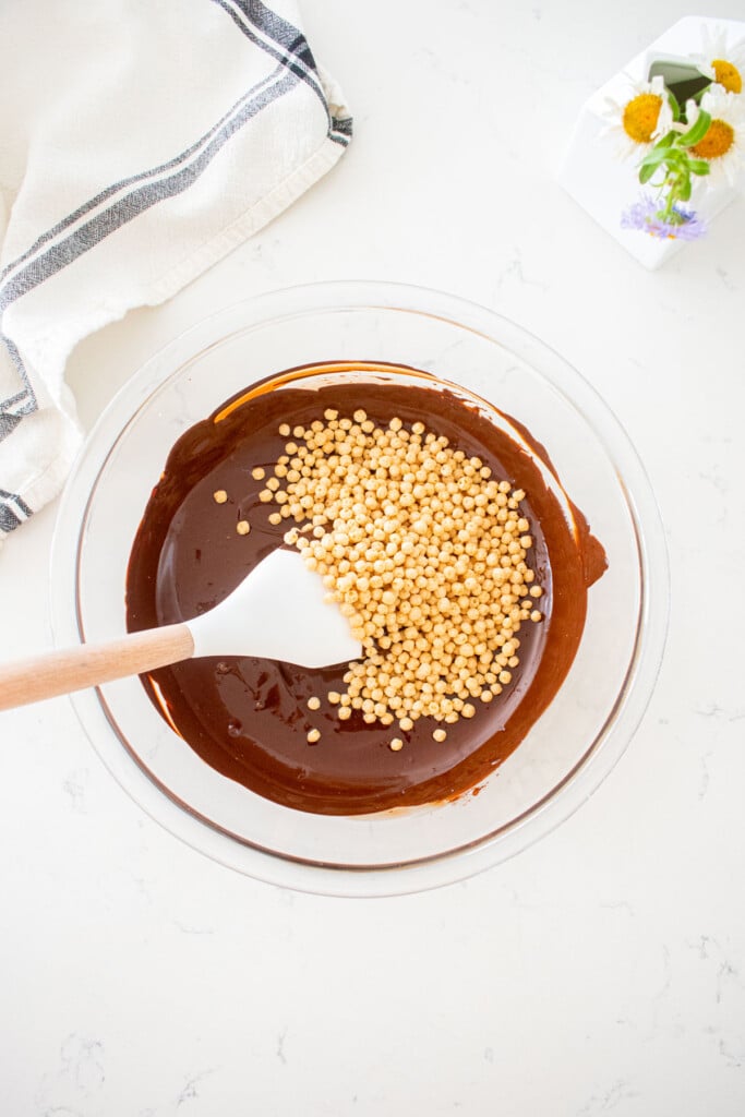 glass bowl with melted chocolate, puffed quinoa and a spatula on a marble counter with a small vase of flowers.