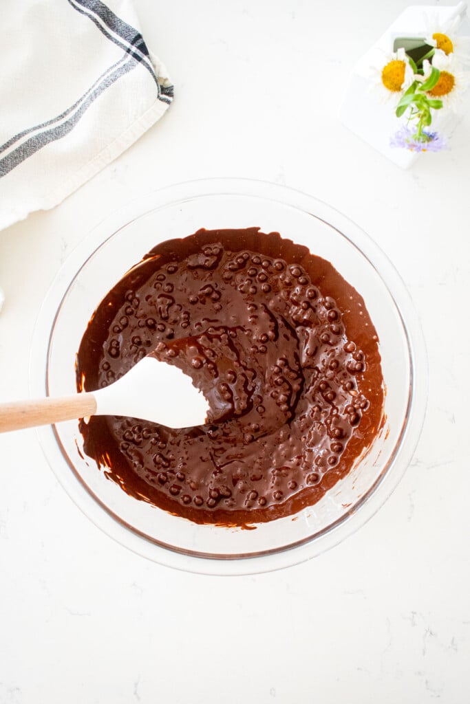 glass bowl of melted chocolate with mixed in puffed quinoa on a marble counter with a small vase of flowers.