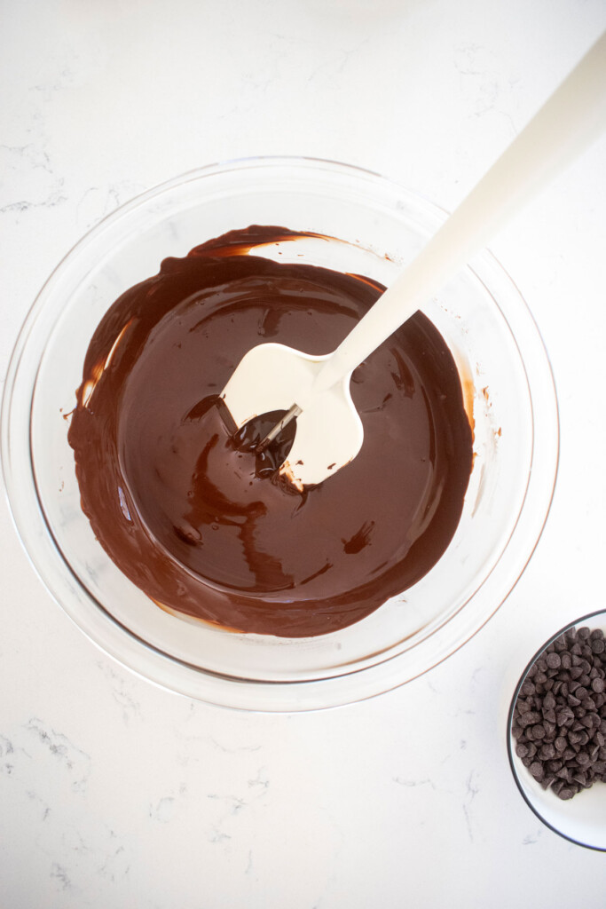 melted dark chocolate in a glass mixing bowl on a white marble counter with a chocolate thermometer in the bowl.