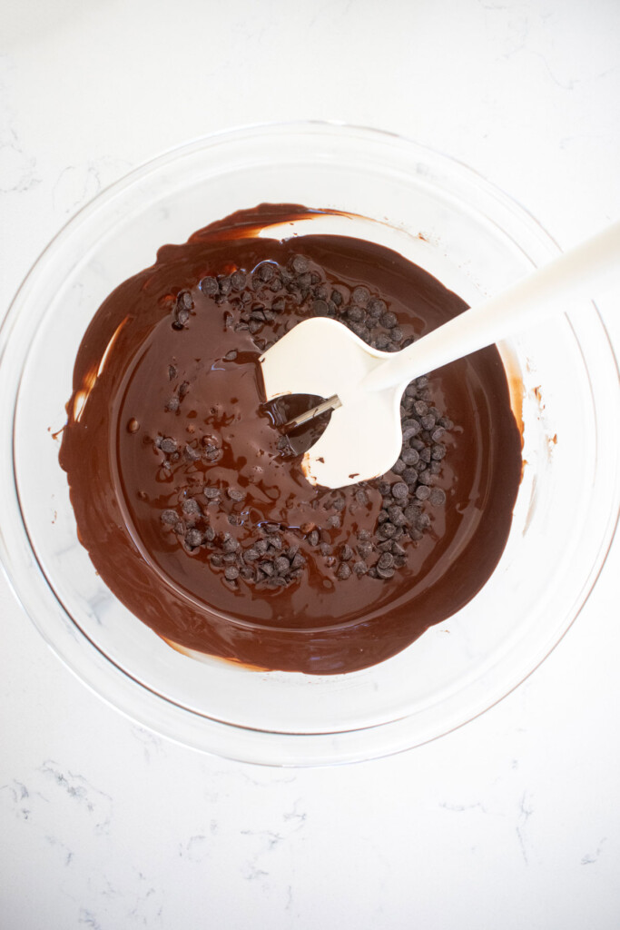 melted dark chocolate in a glass mixing bowl on a white marble counter with a chocolate thermometer in the bowl.