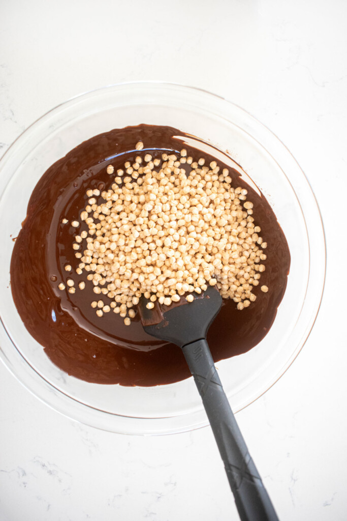 melted dark chocolate in a glass mixing bowl with puffed quinoa on a white marble counter.