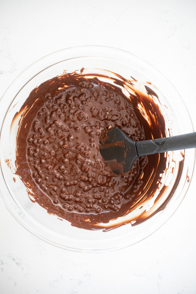 melted dark chocolate in a glass mixing bowl with puffed quinoa stirred into it on a white marble counter.