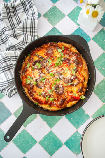 pan pizza in a cast iron skillet with fresh basil and parmesan on top on a green tile table with fresh flowers.