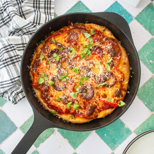 pan pizza in a cast iron skillet with fresh basil and parmesan on top on a green tile table with fresh flowers.