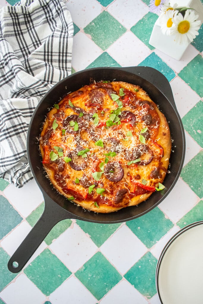 pan pizza in a cast iron skillet with fresh basil and parmesan on top on a green tile table with fresh flowers.