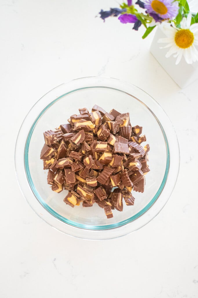 glass mixing bowl of chopped milk chocolate peanut butter cups on a marble counter.