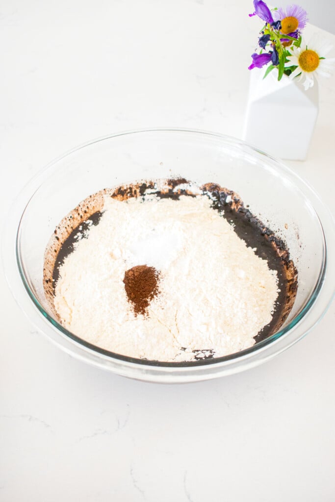dry ingredients added to wet ingredients for brownies in a glass mixing bowl on a marble counter.