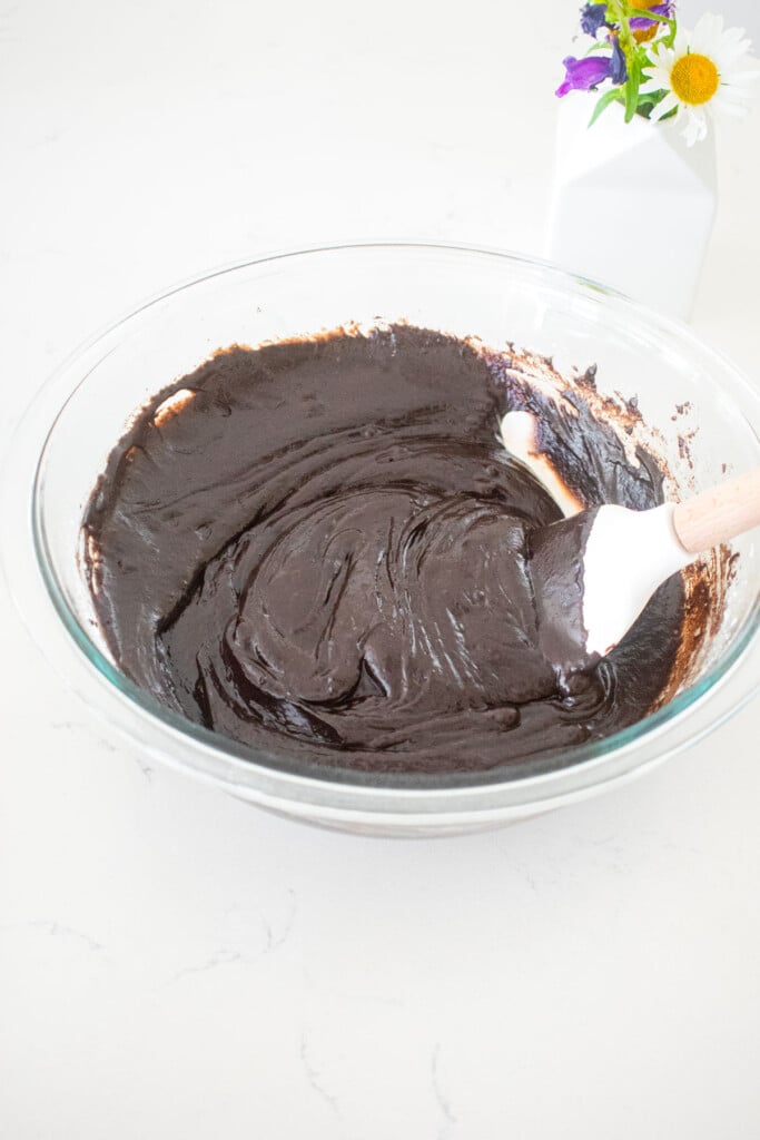 dark chocolate brownie batter in a glass mixing bowl with a spatula on a marble counter.