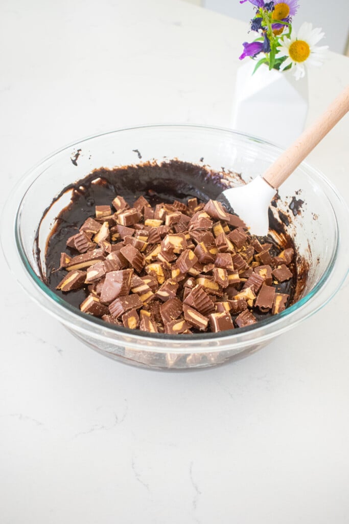 chopped milk chocolate peanut butter cups added to brownie batter in a glass mixing bowl on a marble counter.