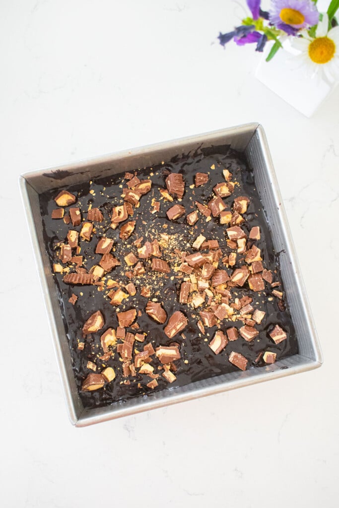 peanut butter cup brownie batter in a square baking pan on a marble counter with more chopped peanut butter cups on top.