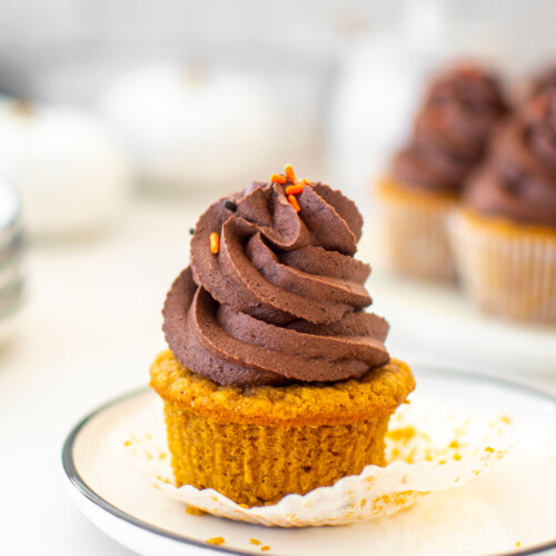 vegan pumpkin cupcake with chocolate frosting unwrapped on a white plate on a marble counter.