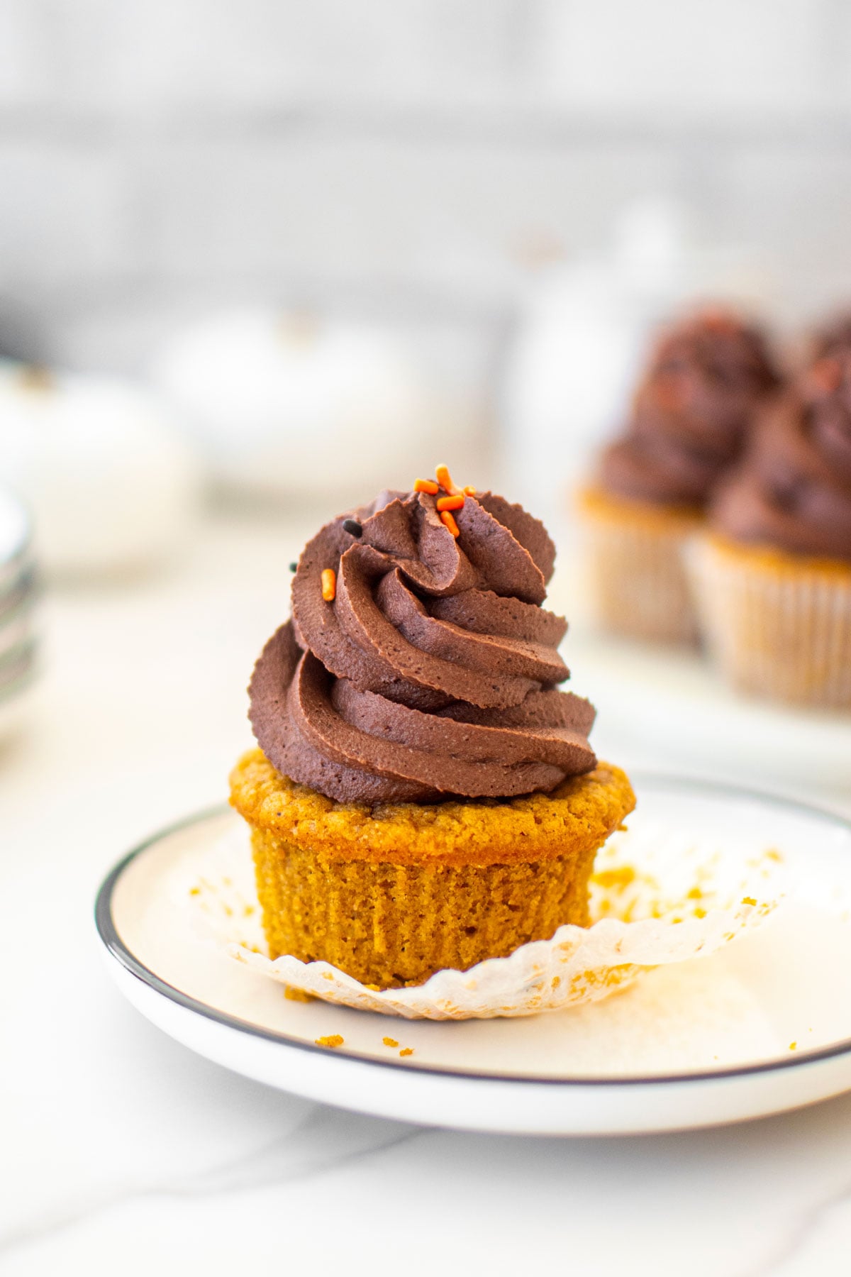 vegan pumpkin cupcake with chocolate frosting unwrapped on a white plate on a marble counter.