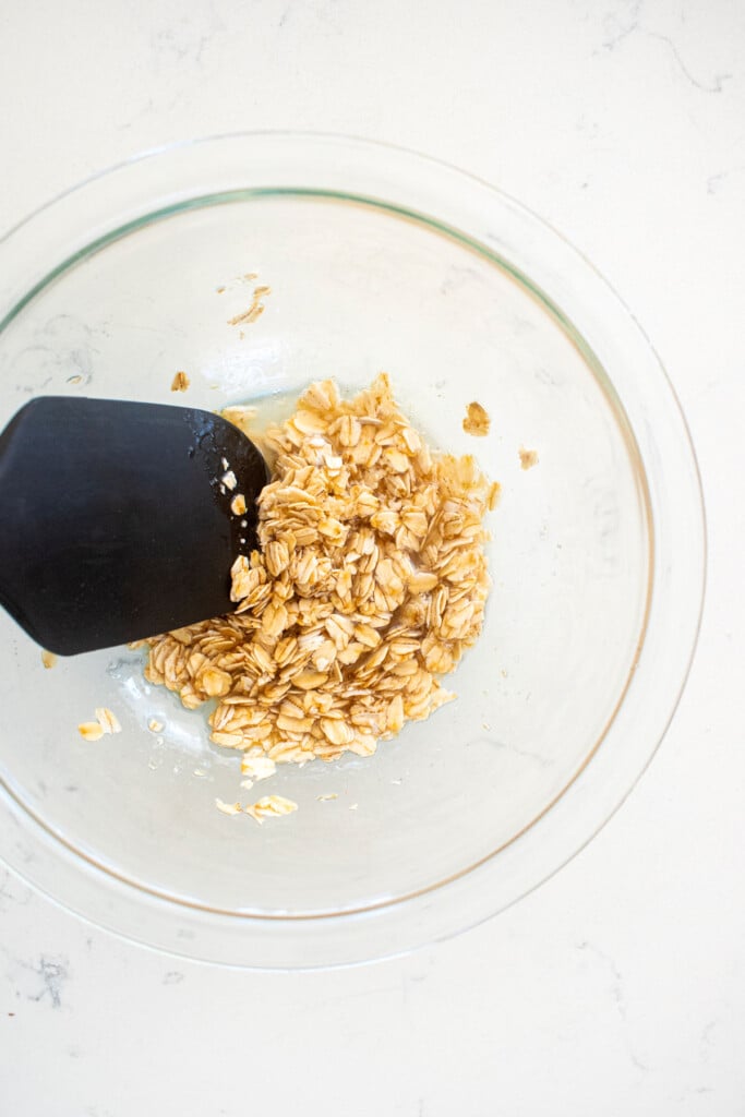 coconut oil, honey and oats mixed in a glass mixing bowl on a white marble countertop.