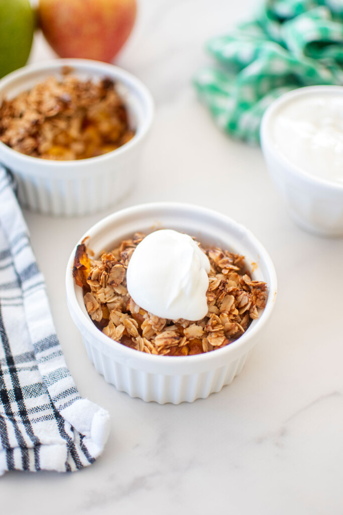 apple crisp for dogs in a small white ramekin with a dollop of greek yogurt on a marble countertop.