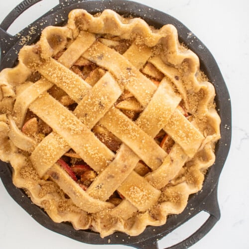 brown butter apple pie with a lattice pie crust top and brushed with honey and turbinado sugar in a cast iron pie pan on a white marble counter.