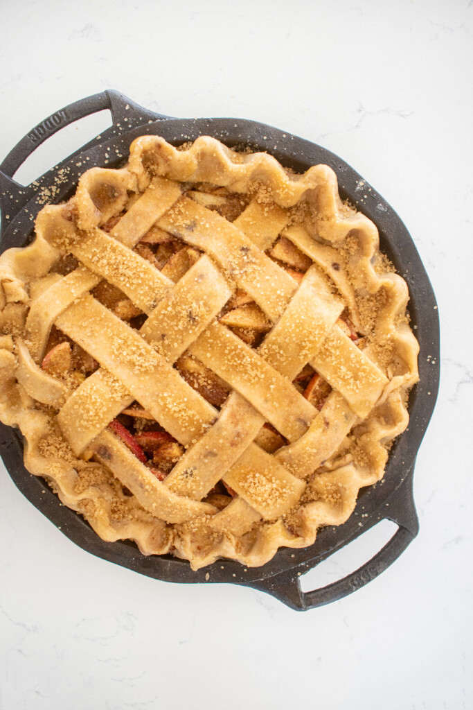 brown butter apple pie with a lattice pie crust top and brushed with honey and turbinado sugar in a cast iron pie pan on a white marble counter.