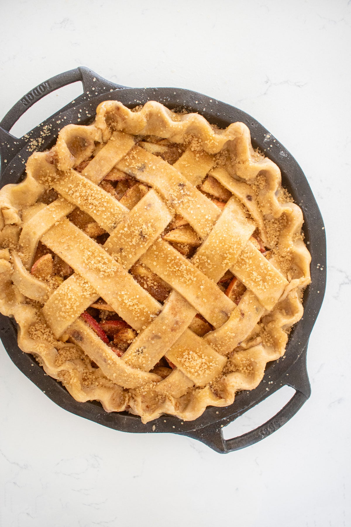 brown butter apple pie with a lattice pie crust top and brushed with honey and turbinado sugar in a cast iron pie pan on a white marble counter.