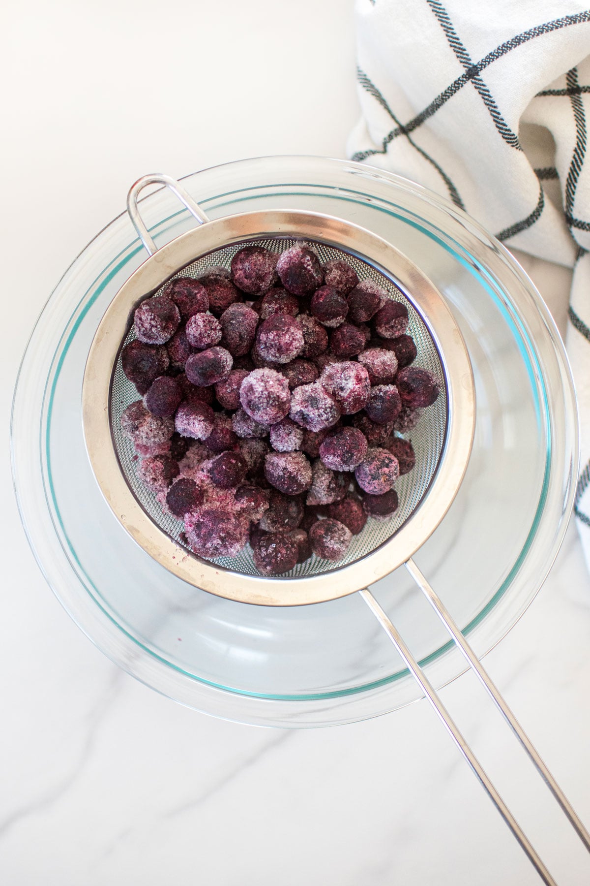 frozen blueberries in a strainer in a bowl on a white marble counter.