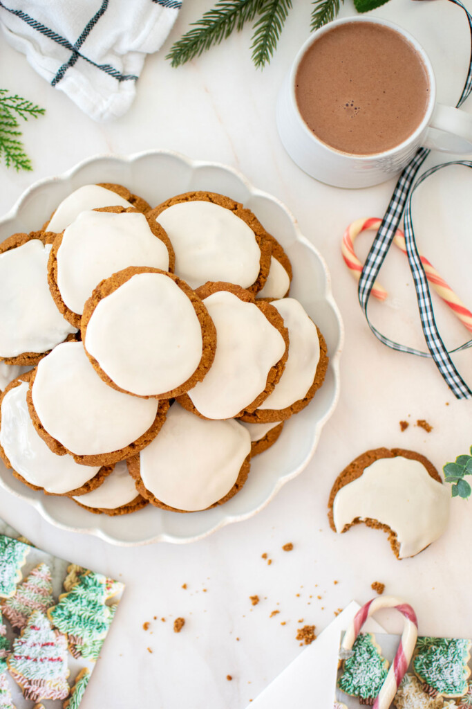 gluten free frosted gingerbread cookies on a plate with a mug of hot cocoa and holiday card on a white marble countertop.