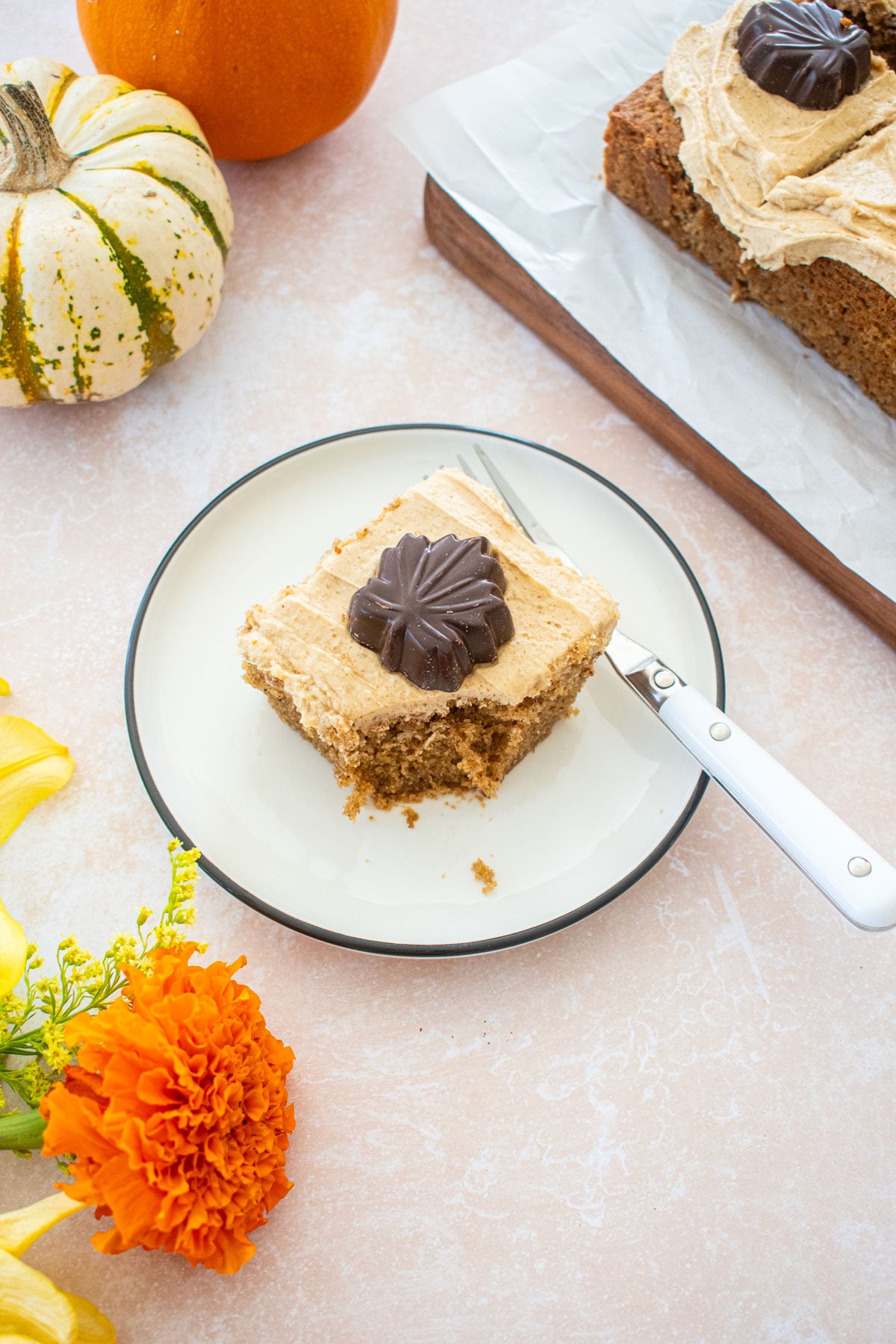 slice of maple spice cake with brown sugar frosting and a chocolate maple leave on a plate with a bite taken out on a table with flowers and tiny pumpkins.