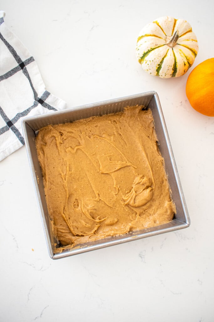 maple spice cake batter in a square cake pan on a marble counter with tiiny pumpkins.
