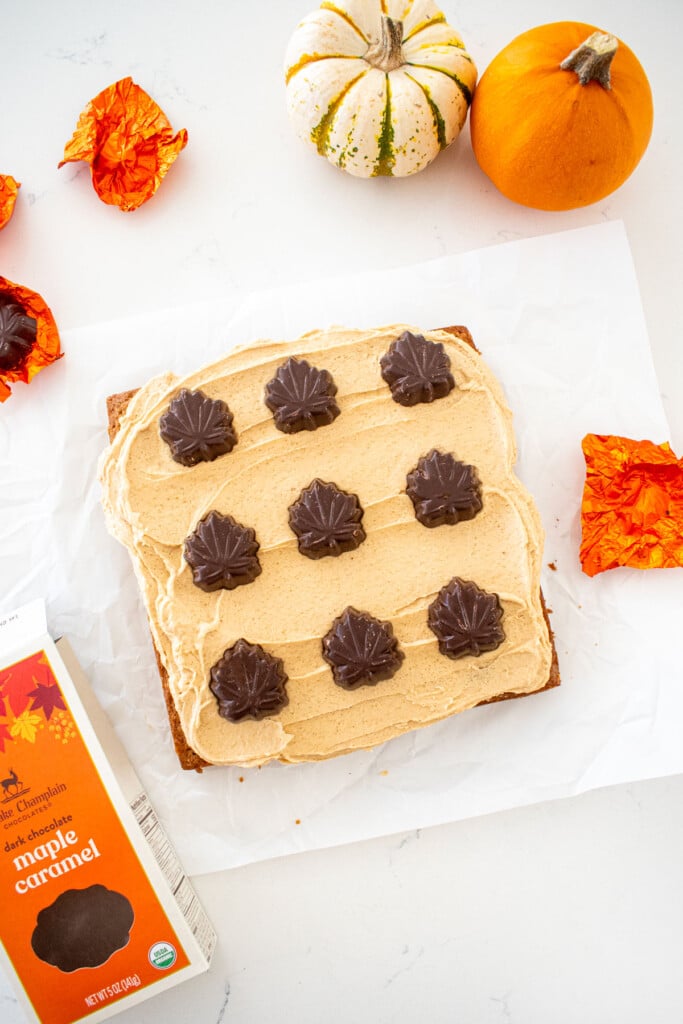square maple spice cake with brown sugar frosting and chocolate maple leaves on top on a marble counter with tiny pumpkins.