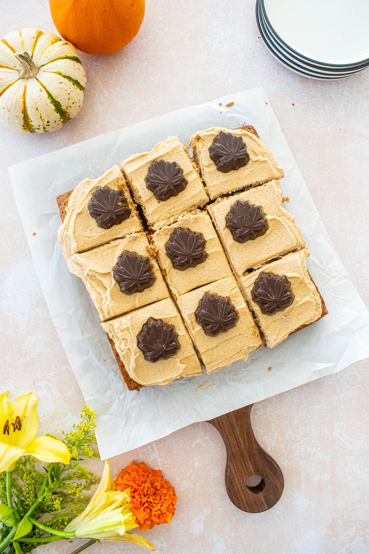 maple spice cake with brown sugar frosting and chocolate maple leaves cut into squares on a table with tiny pumpkins and fresh flowers.
