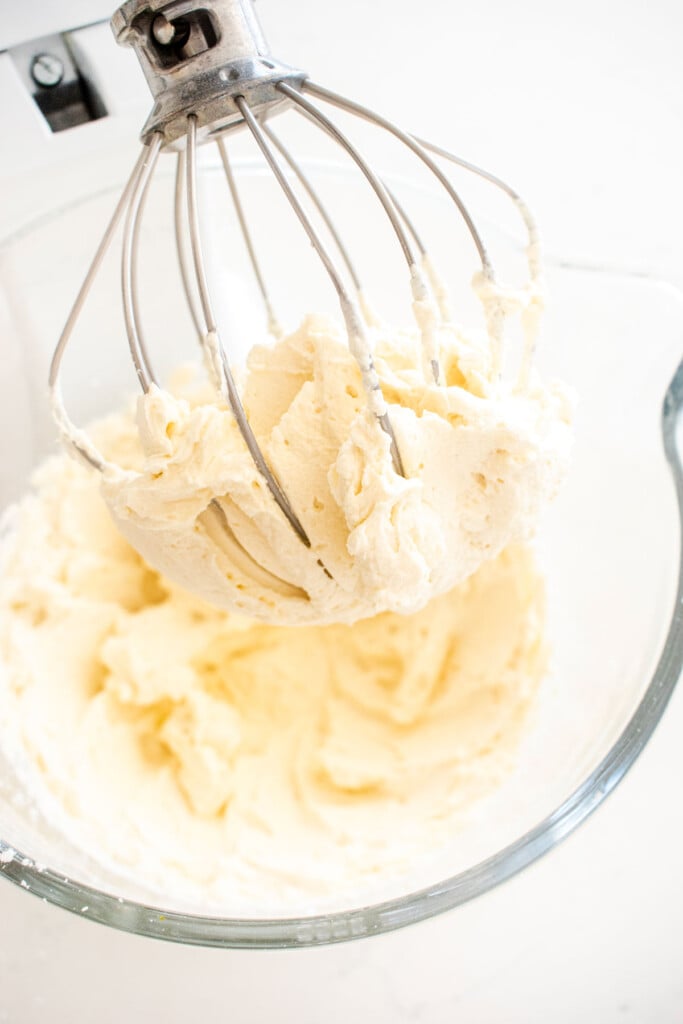 whipped cream frosting in a glass mixing bowl with a whisk on a white marble counter.