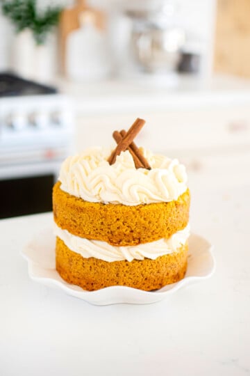 pumpkin pie layer cake with cinnamon sticks on a ruffle cake plate on a marble kitchen countertop.