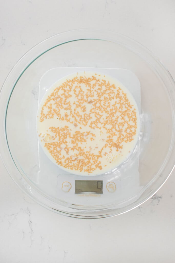 honey, milk and yeast in a glass mixing bowl on a scale on a white marble counter.