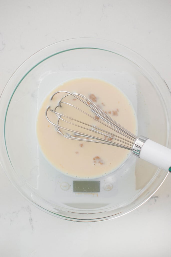 milk, honey and yeast in a glass mixing bowl with a whisk on a scale on a white marble counter.