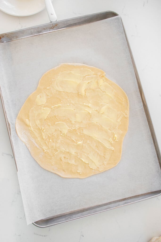 sourdough crescent roll dough being brushed with butter on a baking sheet on a marble counter.