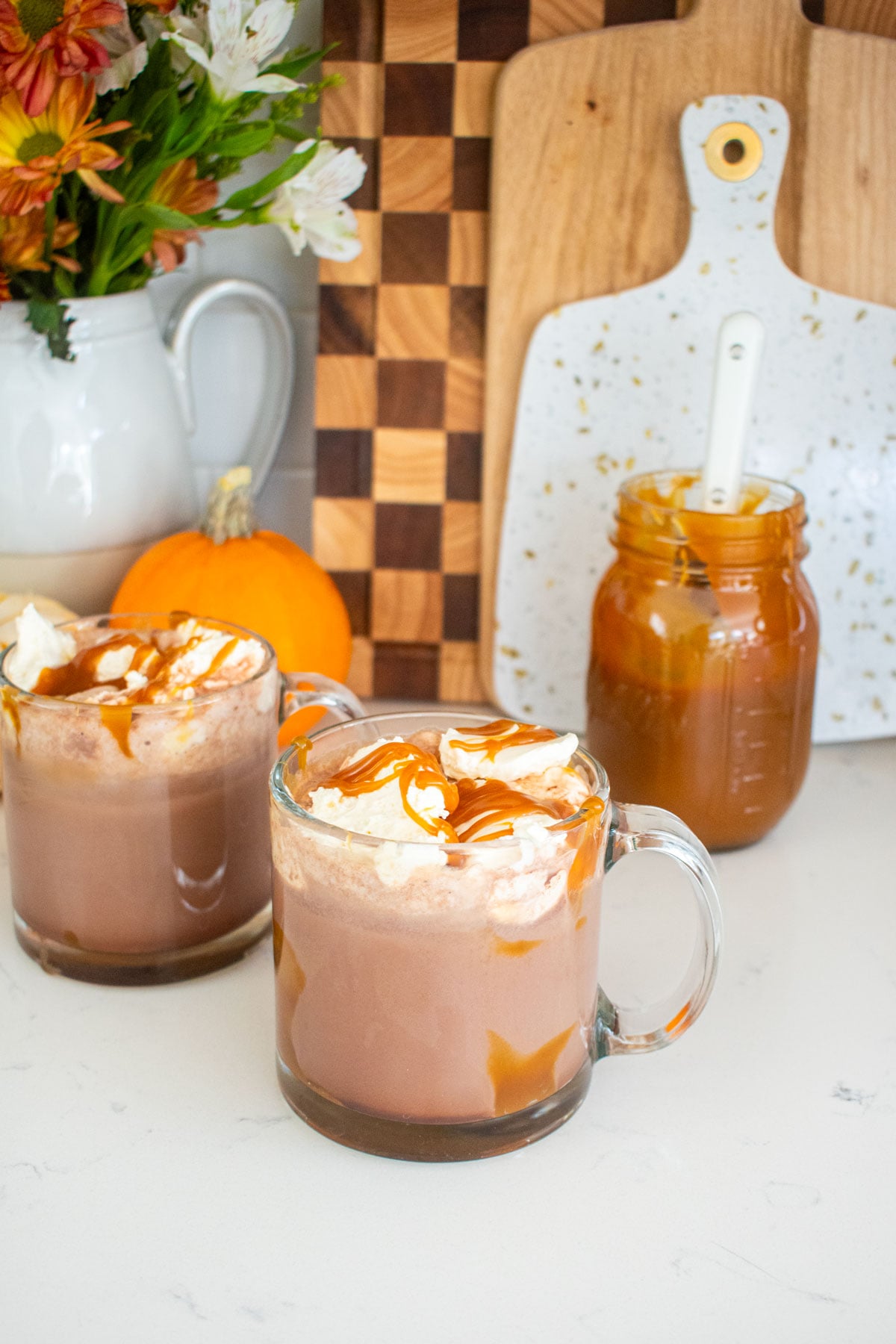 two glass mugs with caramel hot cocoa on a marble counter with whipped cream on top and caramel sauce on top.