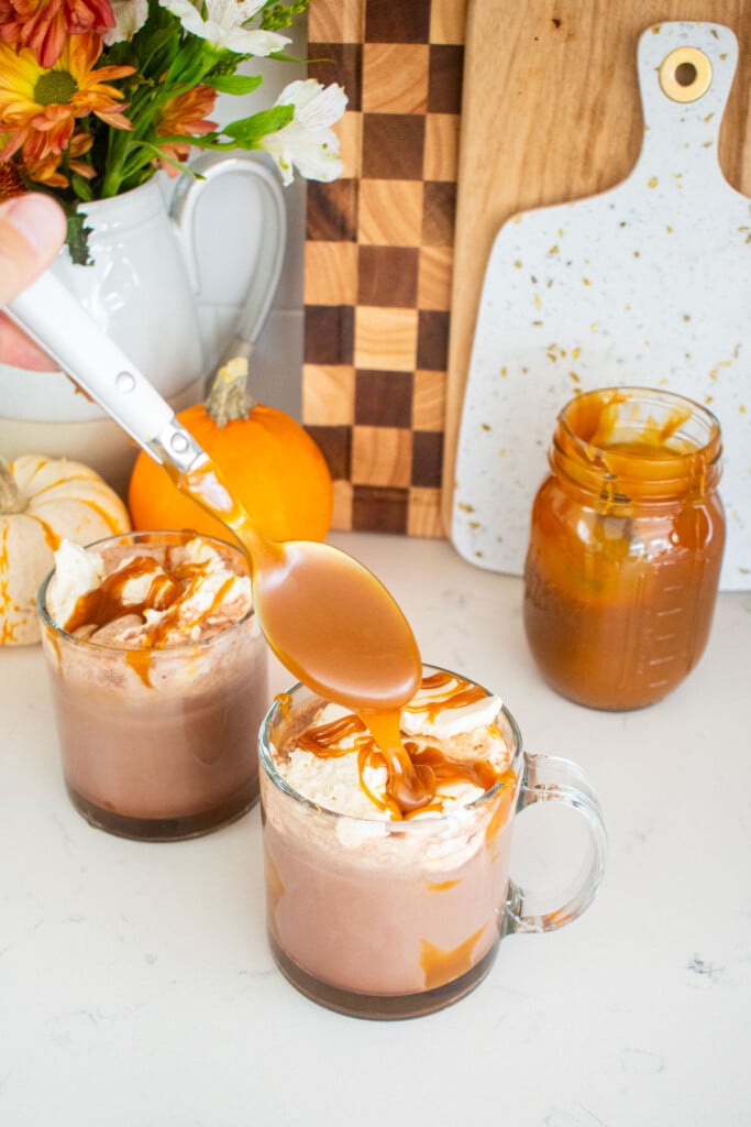 glass mugs with caramel hot cocoa and whipped cream on top with a spoon dropping caramel sauce on top of the whipped cream.