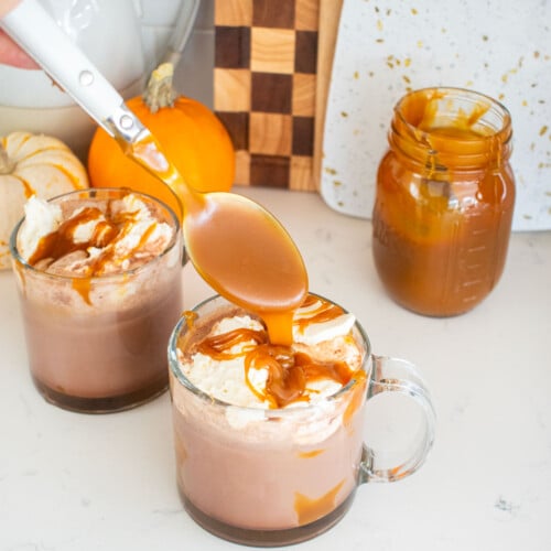 glass mugs with caramel hot cocoa and whipped cream on top with a spoon dropping caramel sauce on top of the whipped cream.