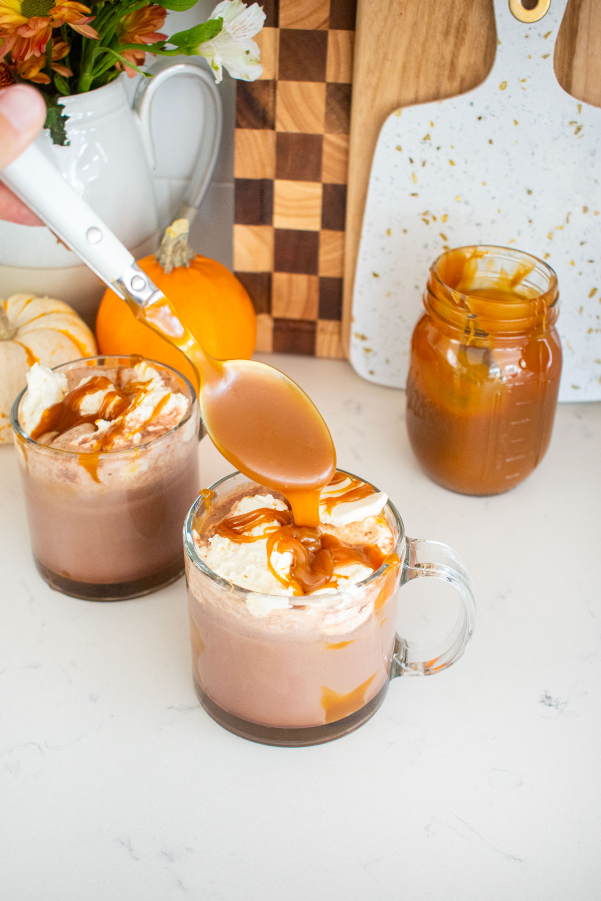 glass mugs with caramel hot cocoa and whipped cream on top with a spoon dropping caramel sauce on top of the whipped cream.