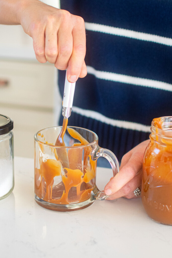 woman smearing caramel sauce into a glass mug at the kitchen counter.