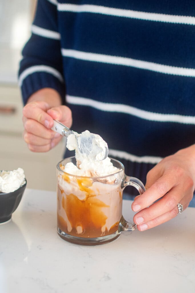 woman adding whipped cream to caramel hot cocoa in a mug at the kitchen counter.