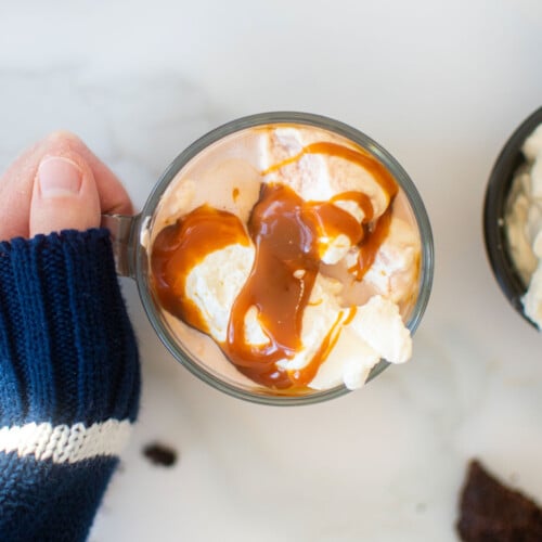 caramel hot cocoa with whipped cream and caramel sauce in a glass mug on a marble counter, woman grabbing with her hand.