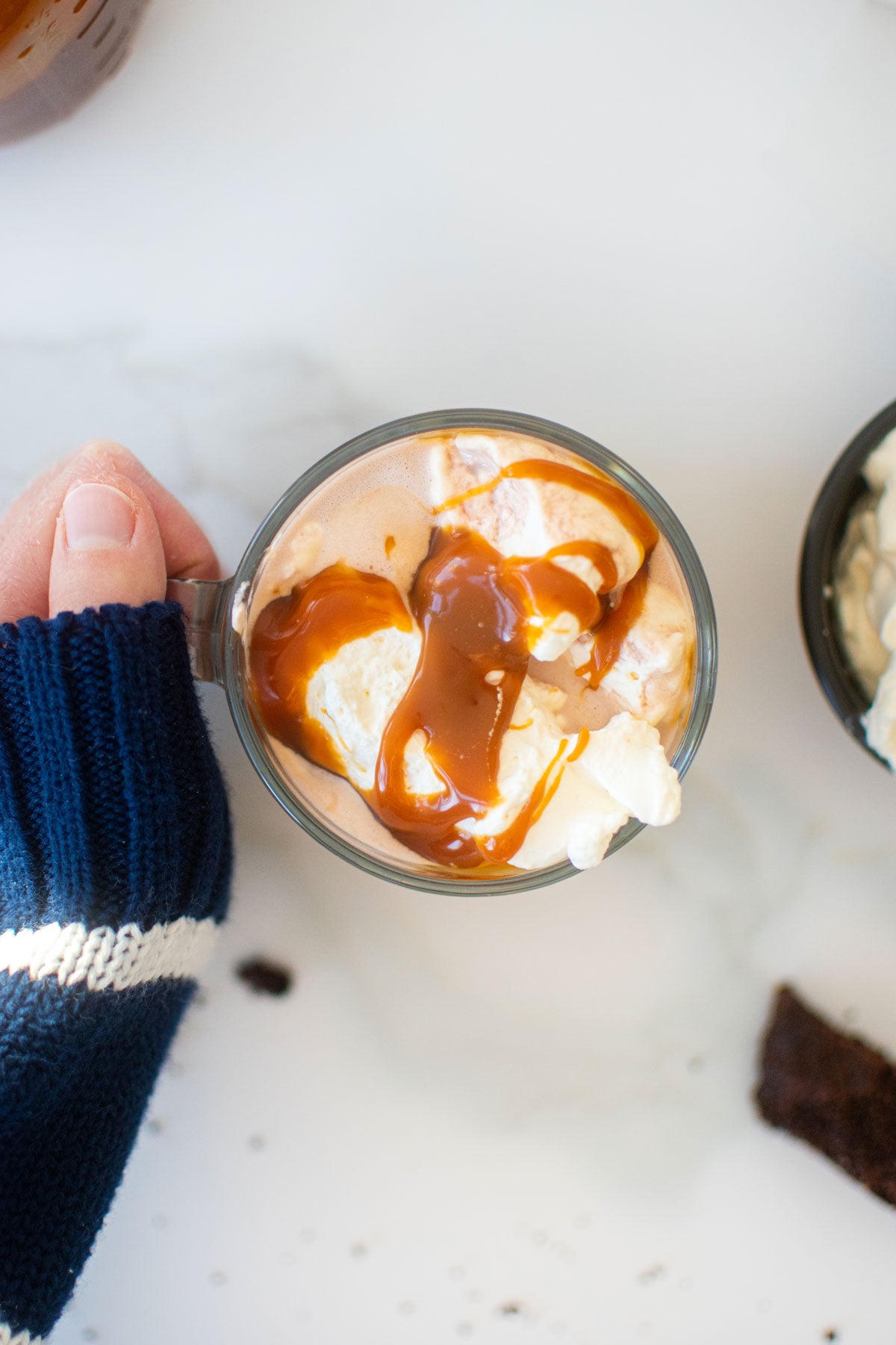 caramel hot cocoa with whipped cream and caramel sauce in a glass mug on a marble counter, woman grabbing with her hand.