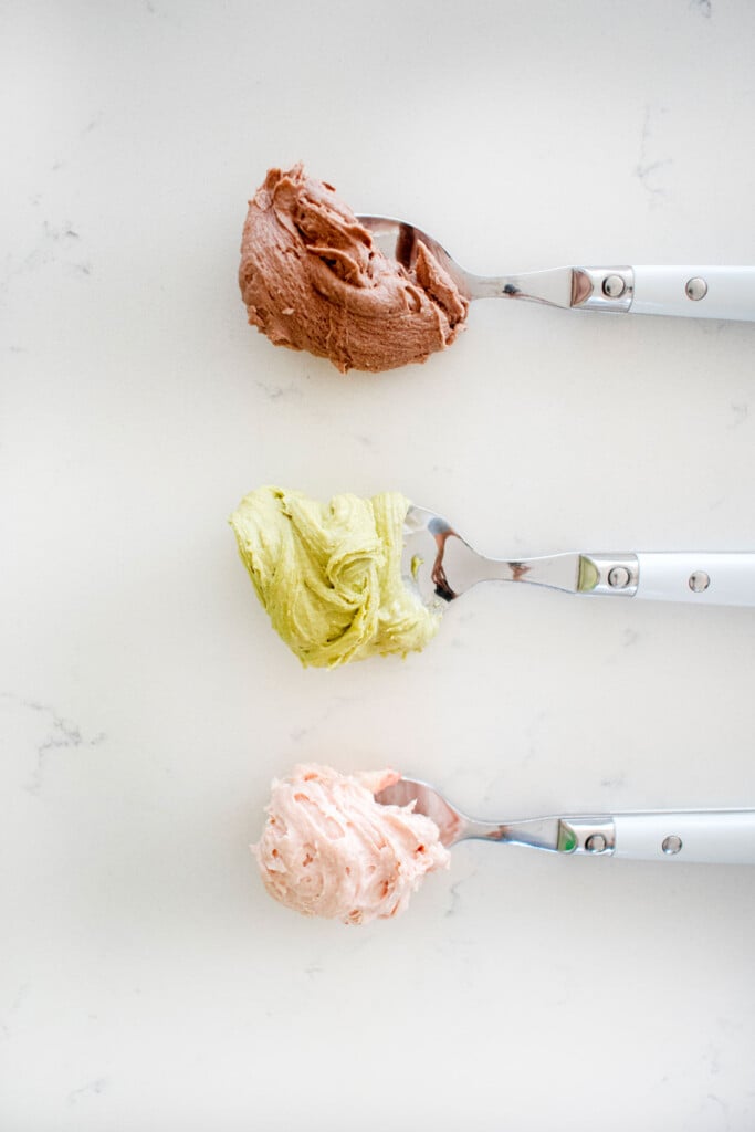 frosting colored brown, green, and link pink on spoons on a white marble counter.