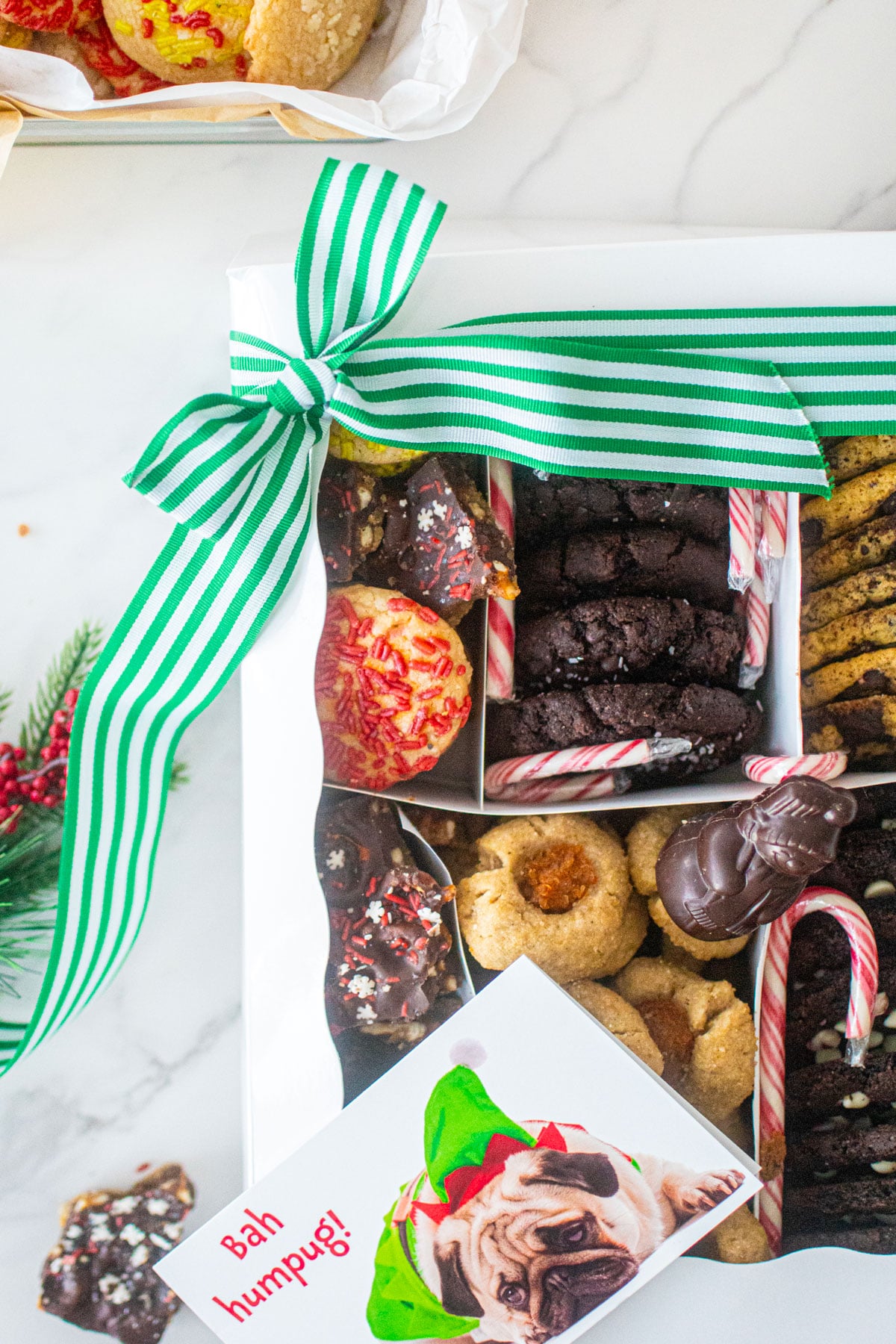 DIY Christmas cookie box on a marble counter tied with a green stripe ribbon and holiday treats around it.