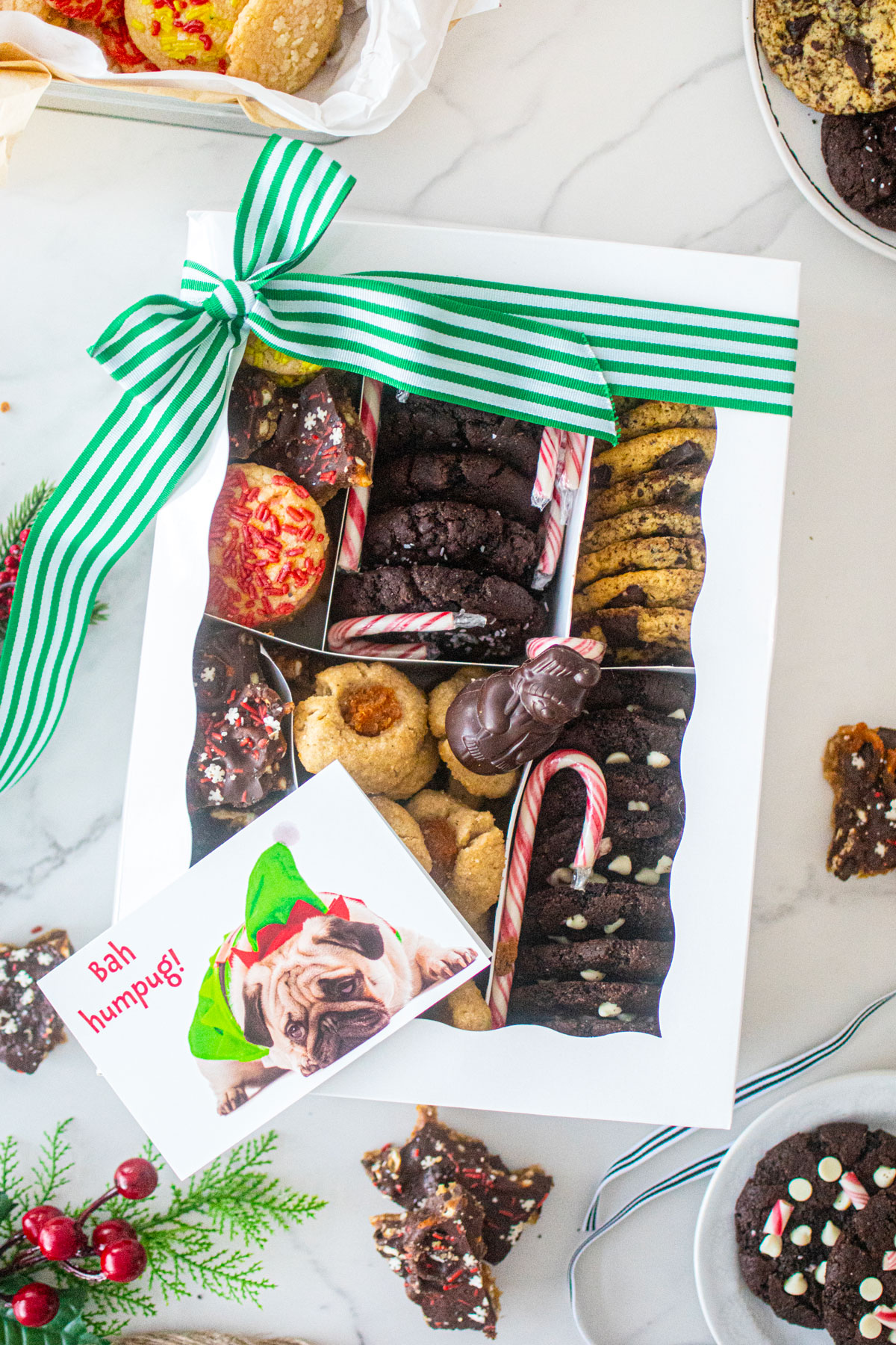DIY Christmas cookie box on a marble counter tied with a green stripe ribbon and holiday treats around it.
