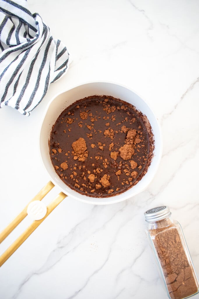 ingredients for gingerbread hot cocoa with bone broth in a small pot on a marble counter with a jar of gingerbread spice and kitchen towel.