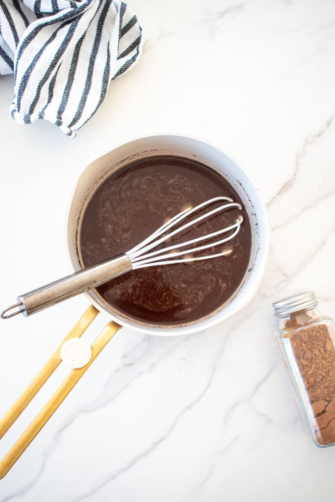 gingerbread hot cocoa with bone broth in a small bot with a whisk on a marble counter with a spice jar and kitchen towel.