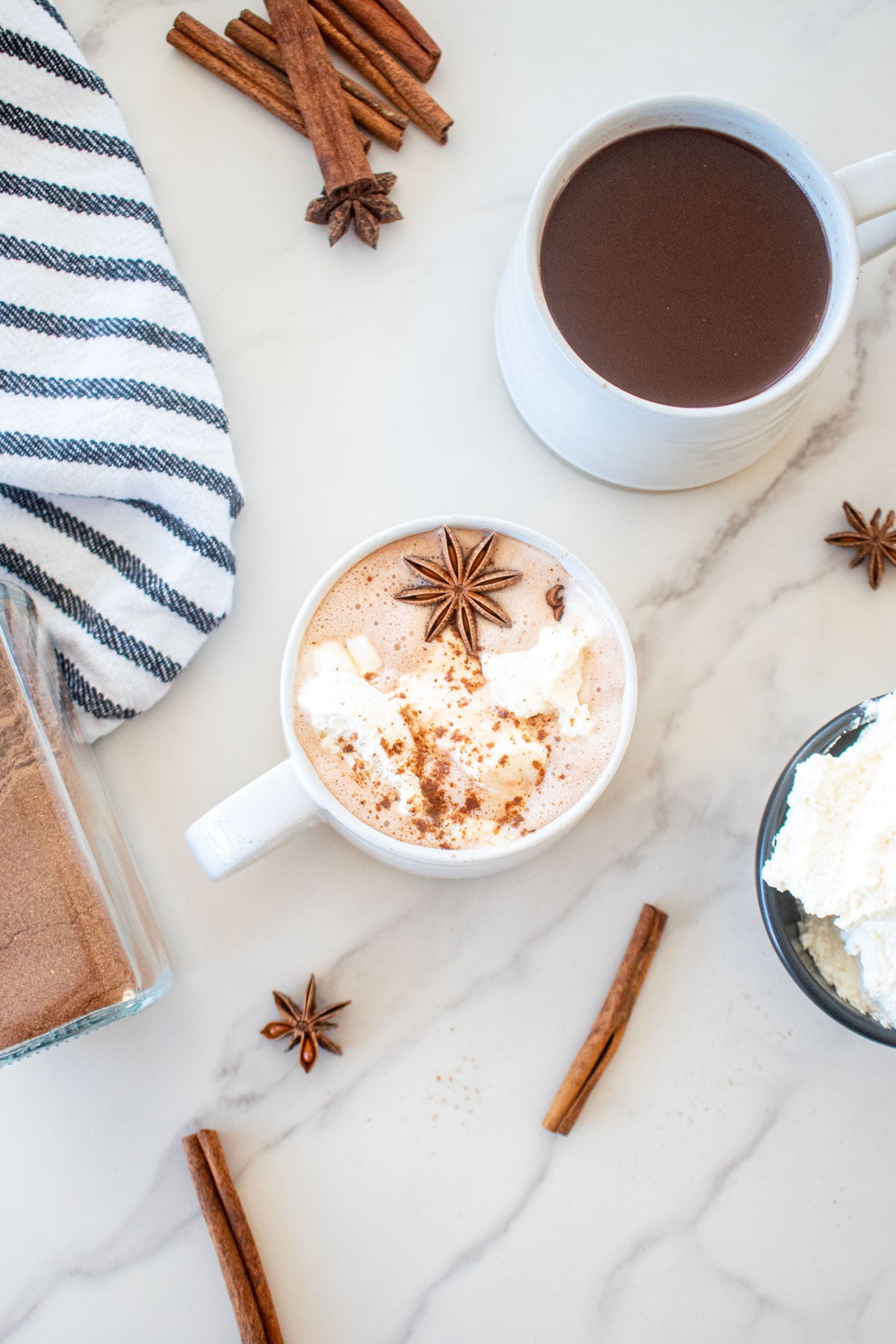 two mugs of gingerbread hot cocoa made with bone broth on a marble counter one has whipped cream, cinnamon stick and star anise on top.