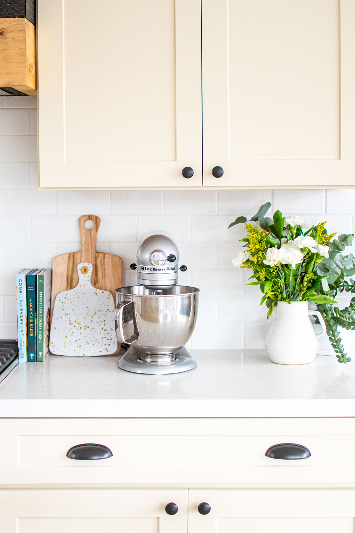 stainless steel stand mixer on a white marble counter with cookbooks, cutting boards and a vase of fresh flowers.
